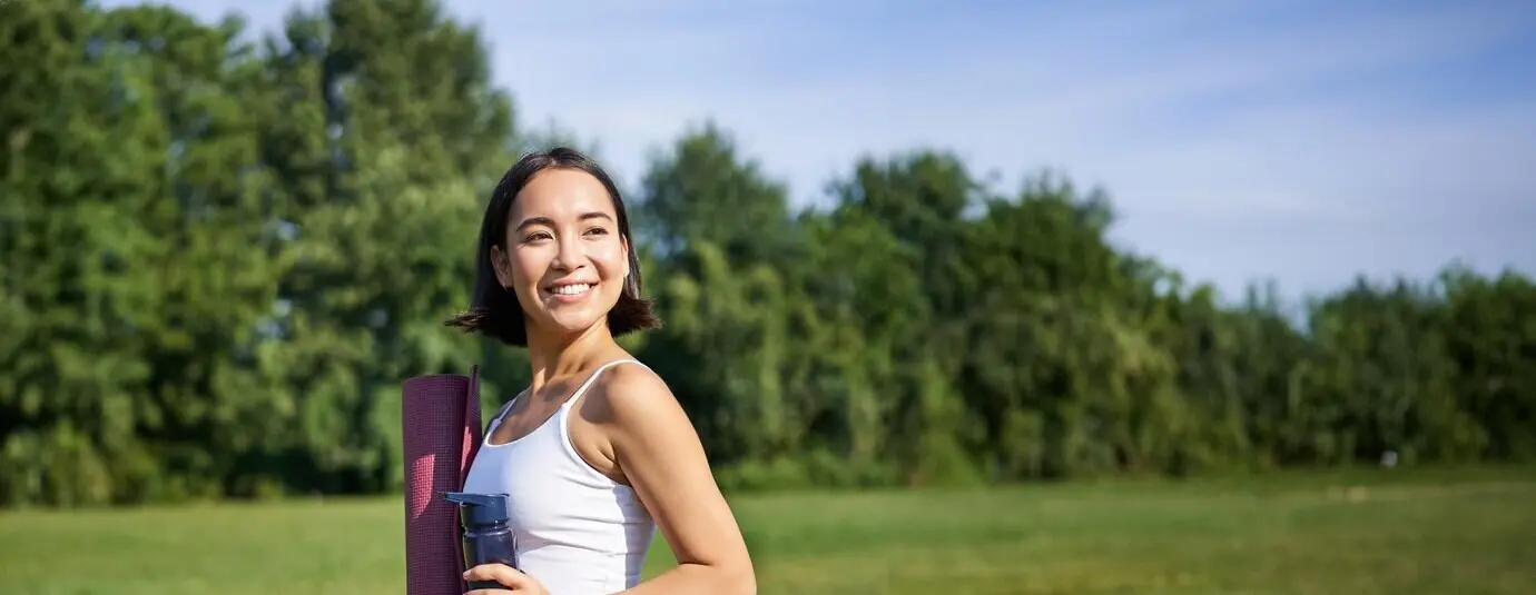 Vertical photo of a fit, healthy Asian woman posing in a park, holding a water bottle and a rubber yoga mat.