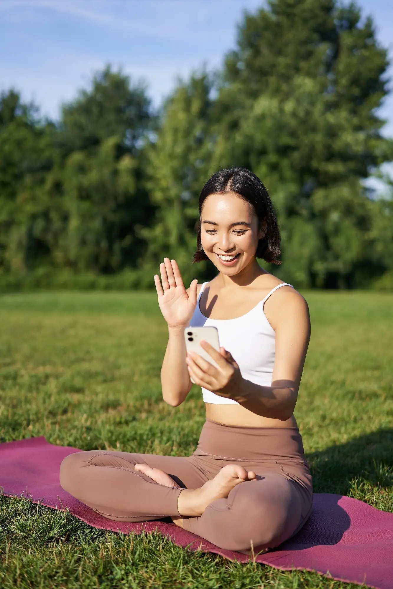 Portrait of an Asian girl seated on a mat in a park, talking to her smartphone while meditating online with yoga instruction.